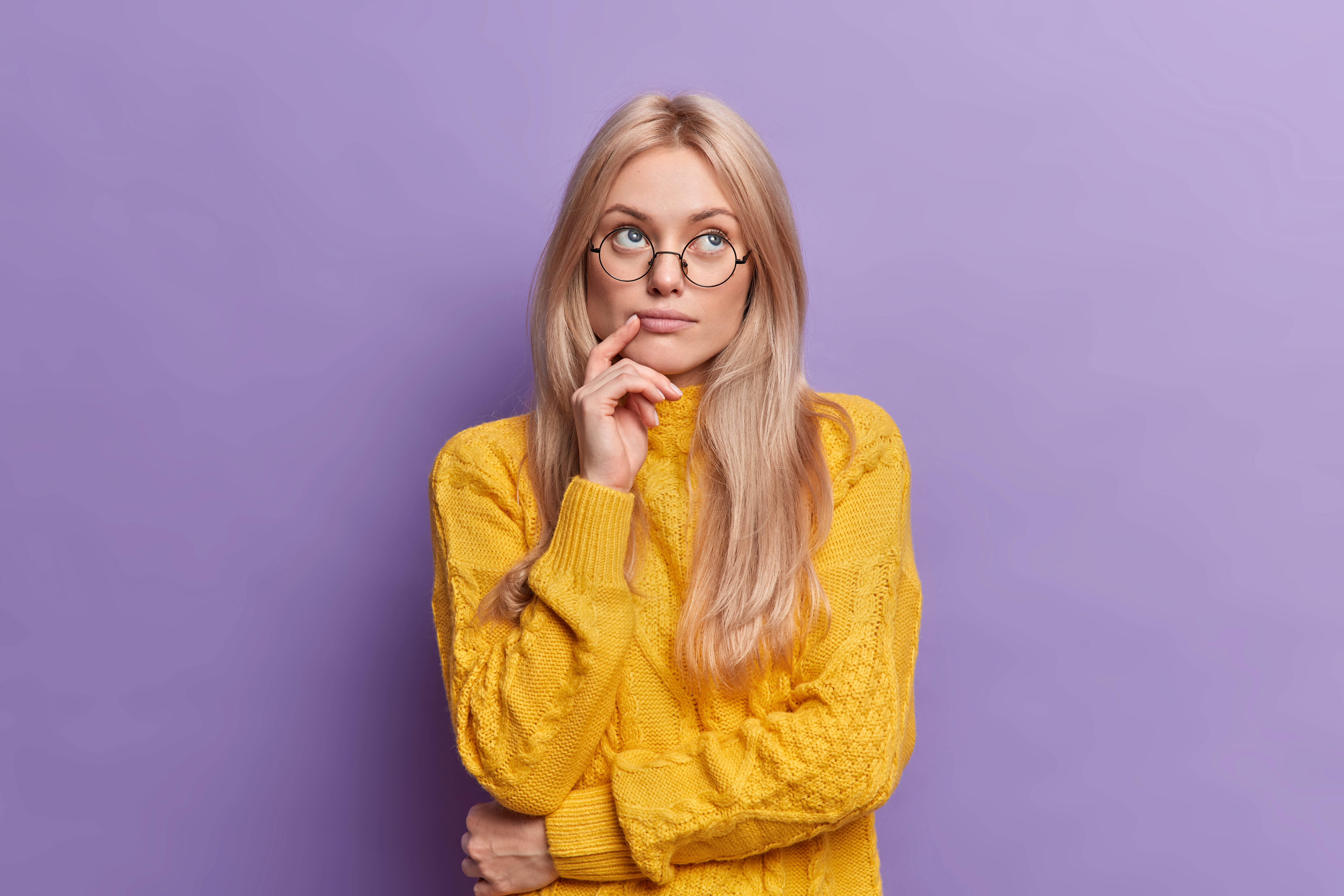Young pretty young woman thinks of ideas concentrated above stands thoughtful and keeps hand on face stands in thoughtful pose wears round glasses yellow sweater isolated over purple background.
